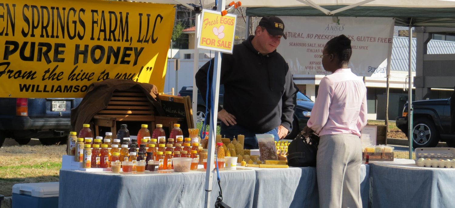 Woman asks questions to a man running a local honey stall in the farmers market. 