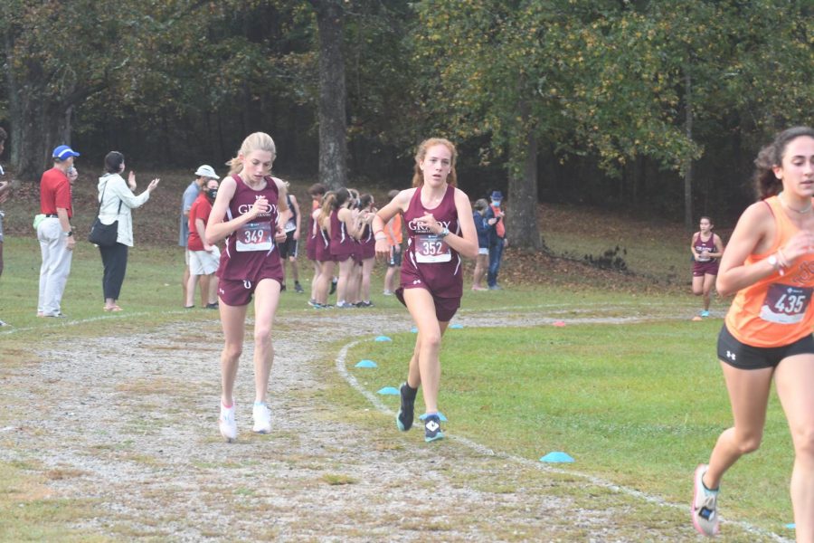 Junior Ellie Spears (left) and sophomore Emilia Weinrobe (right) run together during a race. Head coach Jeff Cramer and the JV girls team cheer on Junior Zoe Chan in the background.