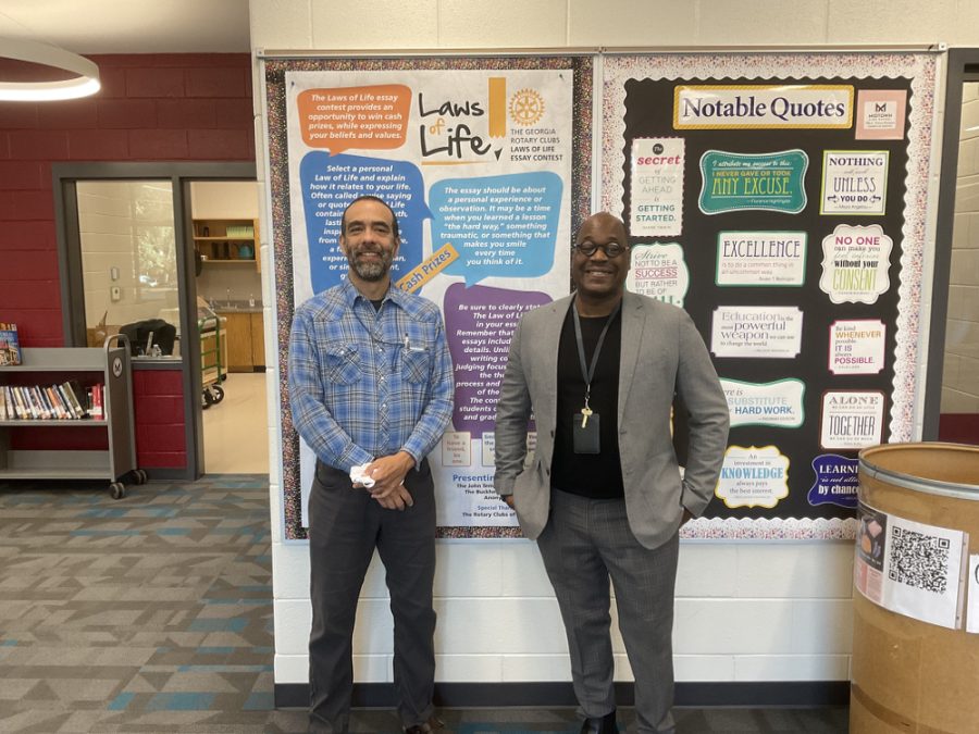 Language Arts teacher Alex Wallace (right) stands with Media Specialist Brian Montero (left) in front of the Laws of Life Essay Contest poster, in the media center.