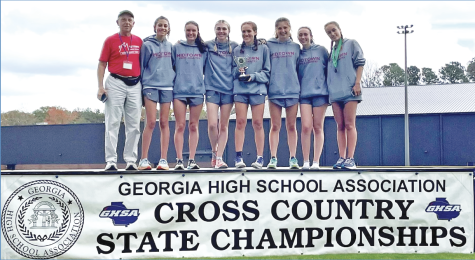 Head Coach Jeff Cramer and the top seven runners celebrate with their second-place trophy, making school history in the state cross country meet. Left to right: Cramer, junior Jameson Knight, senior Sarah Prevost, senior Sally Thompson, senior Emilia Weinrobe,  sophomore  Cate Barton, sophomore Sierra Pape, and 7th-place finisher, junior Cary Schroeder.  